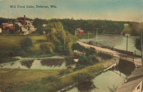 Elevated view of a pond, a stream, and a lake, with a road dividing the pond and stream from the lake. Industrial buildings and a smokestack are in the background, though the area is primarily rural. Caption reads: "Mill Pond Lake, Delavan, Wis."