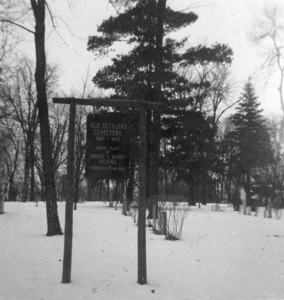 View of a sign to the cemetery that reads: "Old Settler's Cemetery 1837-1875, Burial Place of Samuel * Henry Phoenix, Founders of Delavan 1836." The cemetery is in the background amongst pine trees.