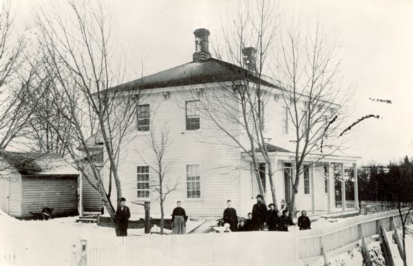 Slightly elevated view of the residence of Casper Hansen, built in 1858. The home has a white picket fence, and a group of people are posing behind the fence in the yard.