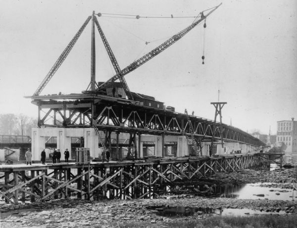 View of the bridge under construction, with a crane atop the structure.  Eight men are standing on scaffolding below the bridge. The Nicolet Paper Company is in the distance to the right.