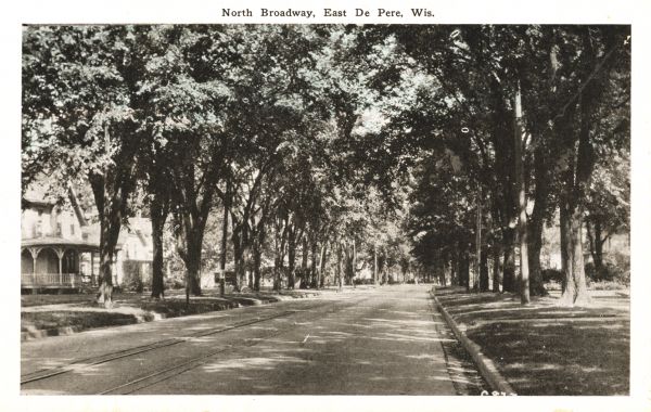 View of residential street with many trees lining the terrace. Rails, possibly trolley rails, run down the center of the street. Caption reads: "North Broadway, East De Pere, Wis."
