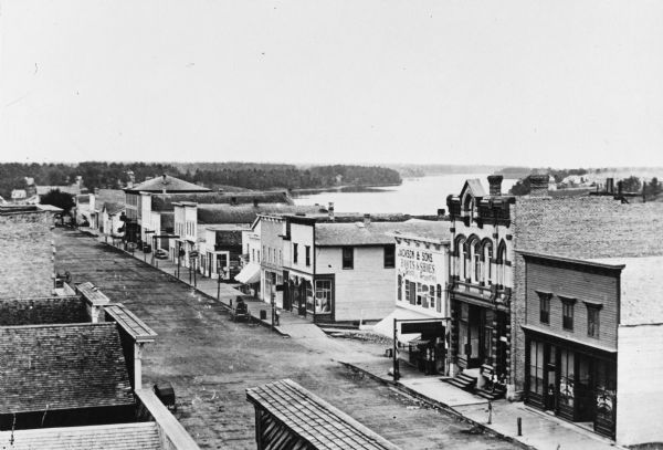Elevated view of a shopping district street near a river, with several businesses lining the street, and two carriages. A building across the street has lettering that reads: "Jackson & Sons Boots & Shoes, Dry Goods and Groceries."