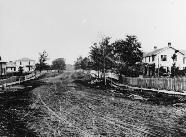 View of dirt road with houses along both sides, and wooden walkways and picket fences along the road.