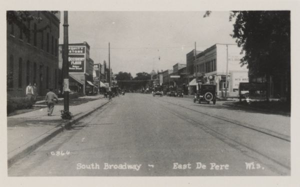 Caption reads: "South Broadway - East De Pere Wis." View down street with pedestrians on the sidewalks and cars traveling down the street.