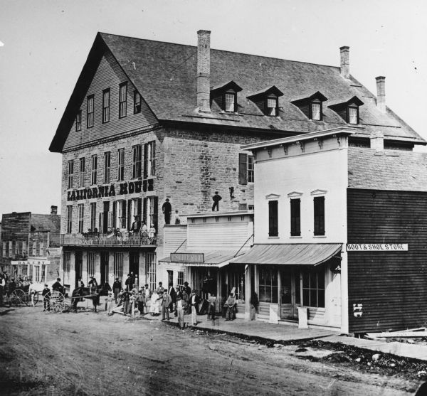 View of the California house next to a store with a sign that reads: "M. Silber & Bro.," and a "Boot & Shoe Store." A bakery is on the left, and a group of people are standing on the sidewalk in front of the buildings. Two horse-drawn carriages are near the bakery.