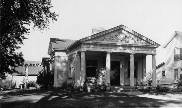 View of front of the building, which is under the shade of a large tree to the left. There is a spinning wheel on the porch. The building has four doric columns and a trapezium or a frieze.