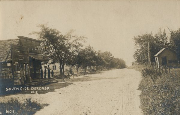 View down a dirt road with a general store on the left, with a sign that reads: "Merchandise". Men and a child are standing on the porch. On the right side is a fence and overgrown grass, and further down a small building.