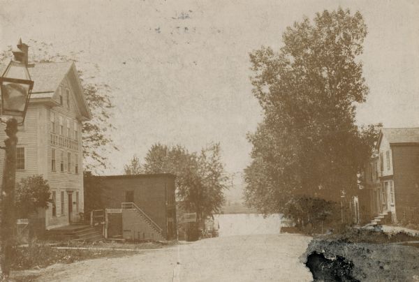 View down road with the Bay State House on the left. Three houses and a barbershop also line the street.  A lamppost is in the left foreground. The Mississippi River is in the distance.