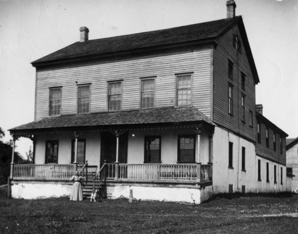 View of the tavern, with a woman in a long dress and a dog on the front lawn. Carriage house, stables, and smoke house are in the rear.  The tavern was built about 1870, and boasted a large dance hall on the second floor and seating for forty people.