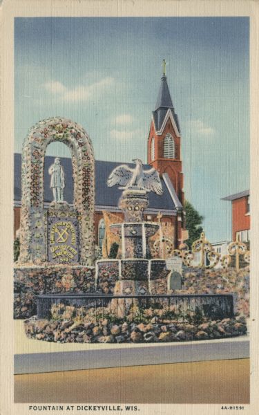 View of fountain with a statue of a bird on top. There is a memorial to Christopher Columbus behind the fountain, and a church with a steeple in the background. Caption reads: "Fountains at Dickeyville, Wis."
