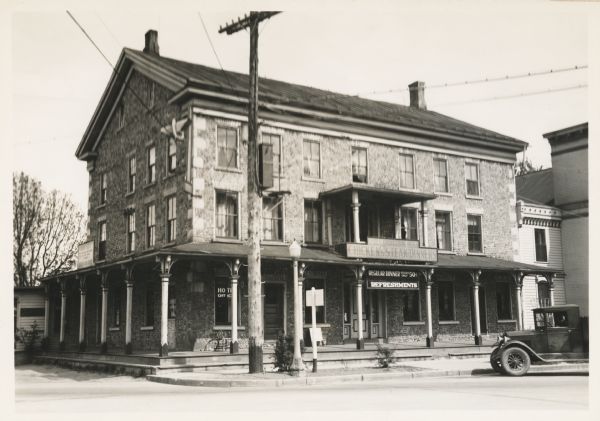 View from street towards the hotel. In front on the terrace is a telephone pole and a lamppost, and on the right is a parked truck. One sign on the hotel reads: "The Cobblestone Hotel, Chicken & Steak Dinners", and another one reads: "Regular Dinner Weekdays Only 50 (cents)." The hotel was formerly the Buena Vista House.