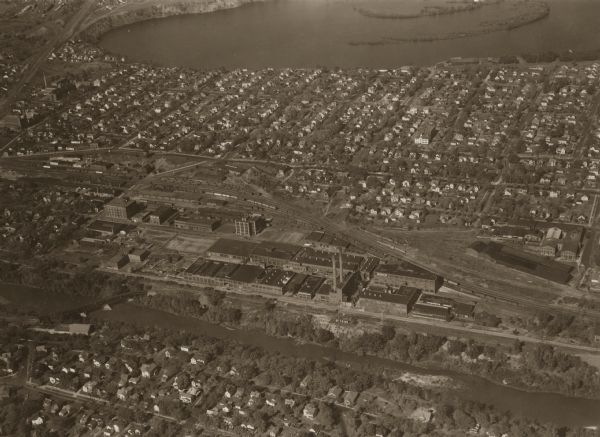 Aerial view of city, with Eau Claire River is in the foreground with Milwaukee railroad bridge at left. Gillette Rubber Co. plant is in center with Omaha railroad tracks and large residential district on the North Side hill. Dells pond or log reservoir at top, with Eddy Street at left.