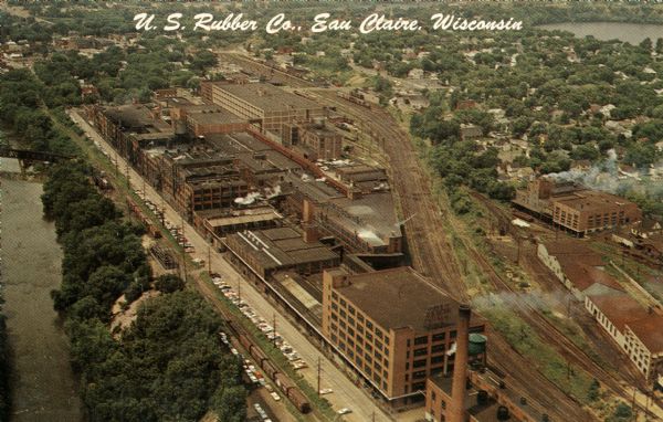 Aerial view of the plant, with railroad tracks along both sides of the property. A river is on the left. Several cars are parked at the plant.  There is another factory on the right. In the distance is a residential area, and a pond or lake is in the upper right. Caption reads: "U.S. Rubber Co., Eau Claire, Wisconsin."