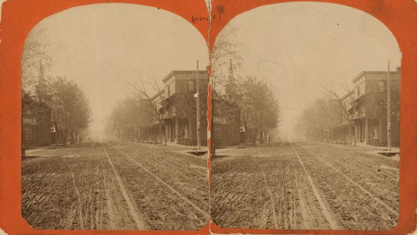 Stereograph view with the Meggett building on the right hand side, and another building along the left hand side. A church's steeple is visible behind the building on the left.