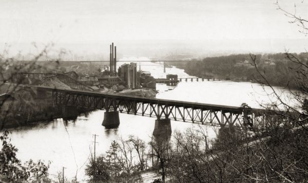 Elevated view of the river with multiple bridges across it, including at least two railroad bridges. There is a factory on the left bank with smokestacks, and what might be a taconite or caliche pile.