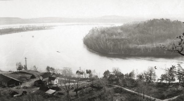 Elevated view of a bow in the river, and a homestead on the shore, with a two-story house and a barn. Two wagons are close to the barn in the lower left corner. A dirt road runs along the river.
