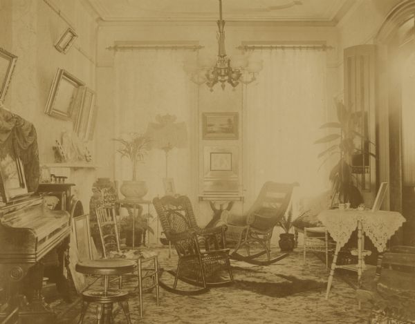 Interior view of a parlor. A chandelier is hanging from the ceiling, and multiple photographs and portraits are displayed on the walls. There is a piano along the left wall, and numerous chairs, including rocking chairs, are in the room which has two large windows on the far wall. Probably the Coffin residence, home of William K. Coffin family.