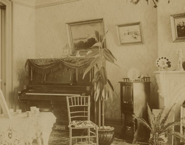 View of the piano in the parlor, with two potted plants on the floor, a chair, and a nightstand. Three framed pictures are hanging on the wall, and a statue stands on the mantle of a fireplace on the right. A small table covered in a doily is in the left foreground. Probably the Coffin residence, home of William K. Coffin family.
