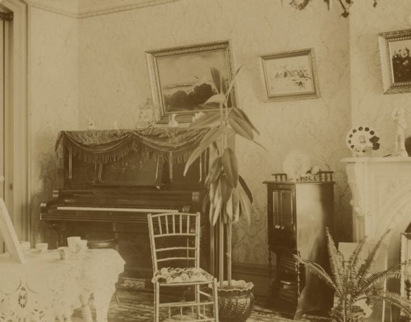 View of the piano in the parlor, with two potted plants on the floor, a chair, and a nightstand. Three framed pictures are hanging on the wall, and a statue stands on the mantle of a fireplace on the right. A small table covered in a doily is in the left foreground. Probably the Coffin residence, home of William K. Coffin family.  