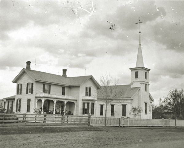 View from road towards the church. The church has a fence running along the front. There are a few people standing on the porch of the parsonage on the left. The church was built in 1859, and the parsonage later. The buildings no longer stand.