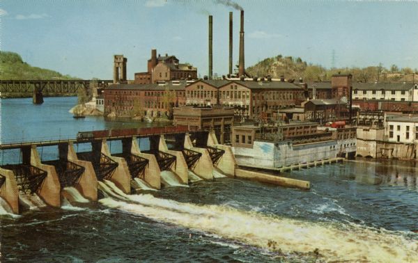 Elevated view of the dam and the power plant. A railroad bridge is in the upper left. Three smokestacks are in the background.