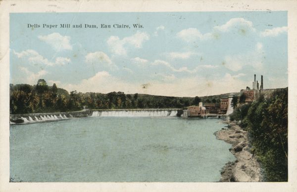 View of the mill and the dam, and the river. The area around the mill is heavily forested. Caption reads: "Dells Paper Mill and Dam, Eau Claire, Wis."