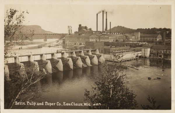 Elevated view of the dam and the mill, with some trees in the foreground. A railroad bridge is in the background on the left, and a rail cart or truck is driving across the dam. Caption reads: "Dells. Pulp and Paper Co., Eau Claire, Wis. "
