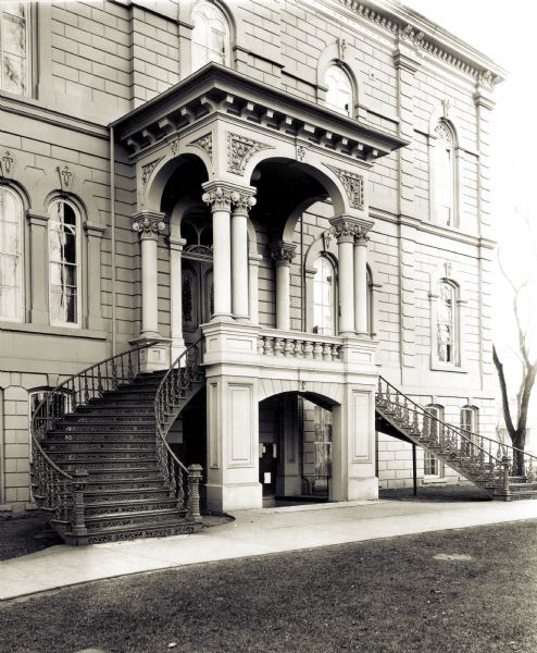 View of the entrance with Corinthian columns, and intricate curved metal stairs and railings flanking the doorway. The sidewalk and lawn are in the foreground.
