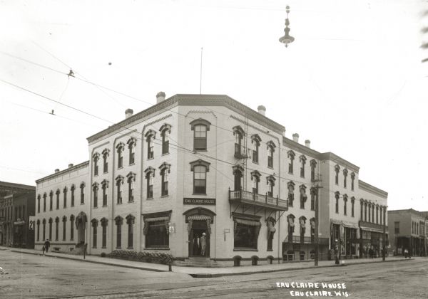 View from street towards the hotel. Two people are in the entry on the corner, and a few people stroll on the sidewalk. Cable car tracks line the road out front, and cable car cables run overhead. Eau Claire House, torn down and replaced by "present"  Eau Claire Hotel. The building shown replaced the first Eau Claire House, a wooden structure, built in 1857, the first hotel of the village.