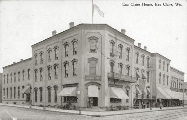 View from street towards the hotel with two people standing under an awning in the entryway at the corner. Cable car tracks are in the foreground. Caption reads: "Eau Claire House, Eau Claire, Wis."