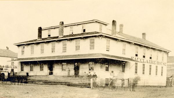 Original part built in 1856, where the present Eau Claire House stands. It was later moved to another site and eventually torn down to make room for the Y.M.C.A. building. The wing at right, with the name of the hotel was not a part of the original building. Blurred movement of people and horse-drawn vehicles in the front of the building.