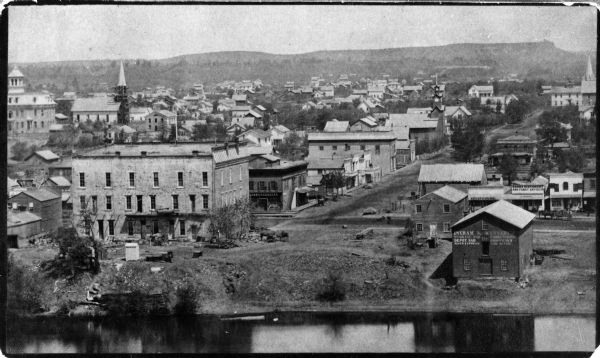 Elevated view of the town, looking northwest. The Chippewa River is in the foreground, with a lot of buildings, an intersection, and distant hills. One building has a sign that reads: "Ingram & Kennedy Storage and Forwarding Depot and Chippewa River Express Office." The three-story building at left center is the newly erected school building that replaced the former school visible at the left rear of the Ingram & Kennedy Storage Depot.
