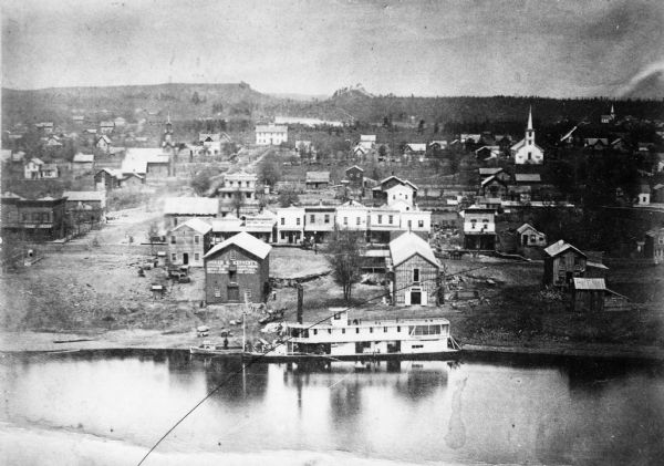 View of the town and Chippewa river, with a steamboat in the center along the riverbank. A smaller boat is next to the steamboat, with a man standing either in the boat or on the shore. The surrounding hills are in the distance. The tallest structure is perhaps a church towards the right.