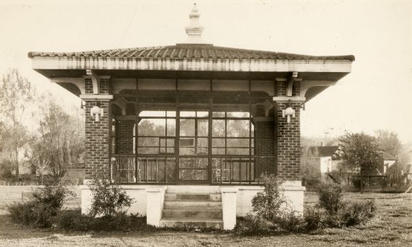 Brick band pavilion, with five steps leading to entrance, shrubbery around the exterior, and houses and trees in the background.