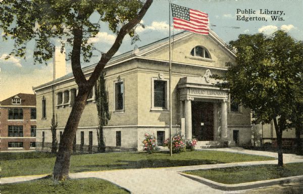 View towards the entrance to the Edgerton Public Library, with a sidewalk in front, and a driveway to the left of the building. An American flag on a flagpole is in the front lawn. Caption reads: "Public Library, Edgerton Wis."