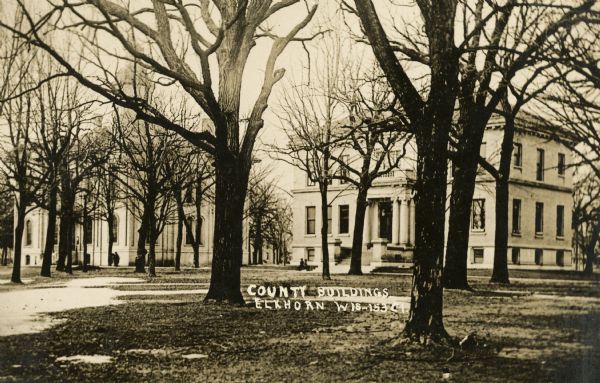 View of buildings through barren trees. There are patches of snow on the ground. Caption reads: "County Buildings, Elkhorn, Wis".