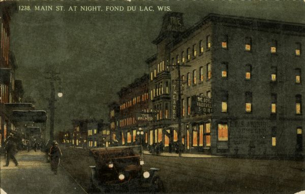Caption reads: "Main St. at Night, Fond du Lac, Wis." A car is parked along the curb in the foreground with its headlights on. A theater is across the street.