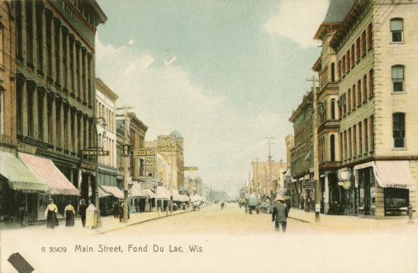 View down Main Street with pedestrians and horse-drawn carriages. Many of the storefronts along the street have awnings. Caption reads: "Main Street, Fond Du Lac, Wis".