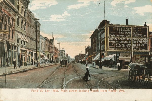 Caption reads: "Fond du Lac, wis Main street looking South from Forest Ave." Horse-drawn carriages and wagons are on the street, as a well as pedestrians on the sidewalk, and a woman in a dress crossing the street.