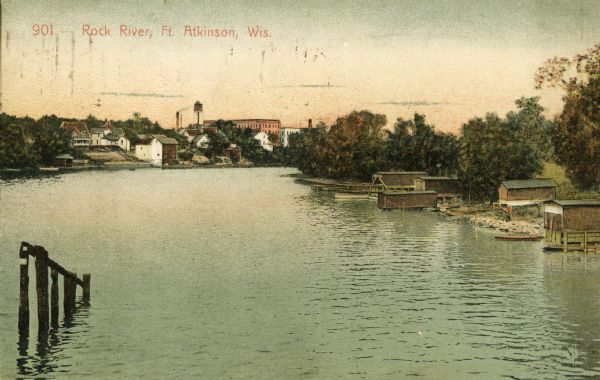 View of the Rock River, with boathouses along the right shoreline, and buildings in the distance on the left at a bend in the river. Pilings are in the foreground. Caption reads: "Rock River, Ft. Atkinson, Wis."