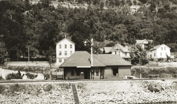 View of a railroad station. In the background are buildings at the base of a bluff.