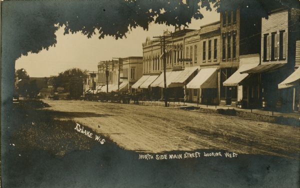 View of Main Street from under the canopy of a tree in the spring or summer. Caption reads: "North Side Main Street Looking West." Across the unpaved street is a row of two or three-story brick buildings with awnings.