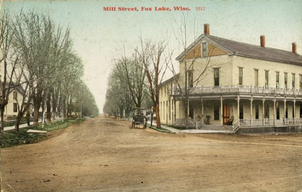 View of an intersection of dirt roads, with a large building, possibly an inn or apartment building on the right corner. A carriage or car is parked near the entrance to the building. To the left is a residential area with houses along a sidewalk lined with trees. Caption reads: "Mill Street, Fox Lake, Wisc."