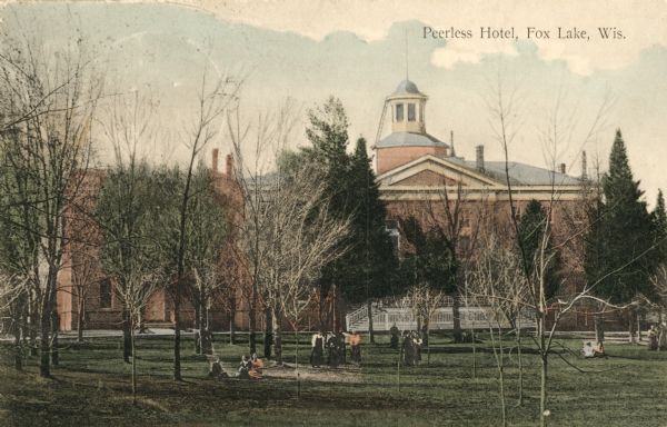 View across lawn, planted with trees, towards the hotel. There are several people either standing or sitting among the trees. Caption reads: "Peerless Hotel, Fox Lake, Wis."