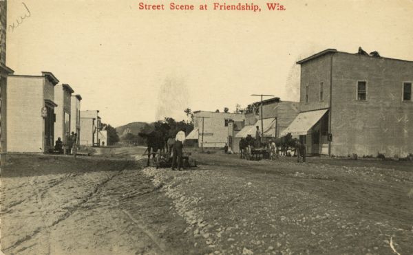 View of a dirt road with storefronts in the background. In the foreground are men with horses and horse-drawn tools. Caption reads: "Street Scene at Friendship, Wis.""