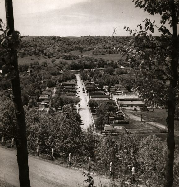 View from hill through two trees over a road with a guard fence, overlooking the town laid out along a main thoroughfare. The area is heavily forested, and tree-covered hills are in the background.