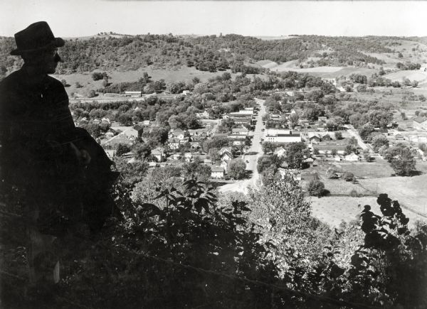 View of Gays Mills from a bluff overlooking the town. A man is sitting on a fence in the left foreground.