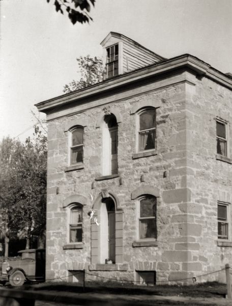 Exterior view of an early stone house. An automobile is parked on the left side of the building.