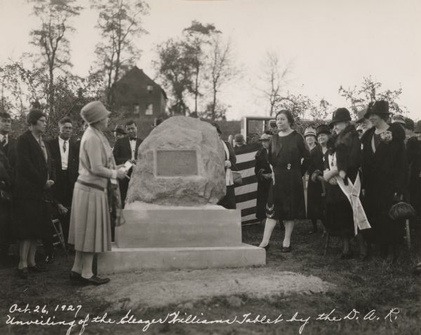 View of a crowd of formally dressed men and women standing around a stone monument with a plaque on it. The edge of what may be a large American flag is visible behind the monument.