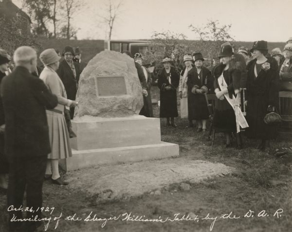 View of a crowd of formally-dressed men and women standing around the plaque mounted on a a large rock.